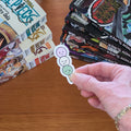 A hand holds a die-cut sticker featuring three dango on a stick (pink, white, and light green) on a wooden table, with manga books in the background.