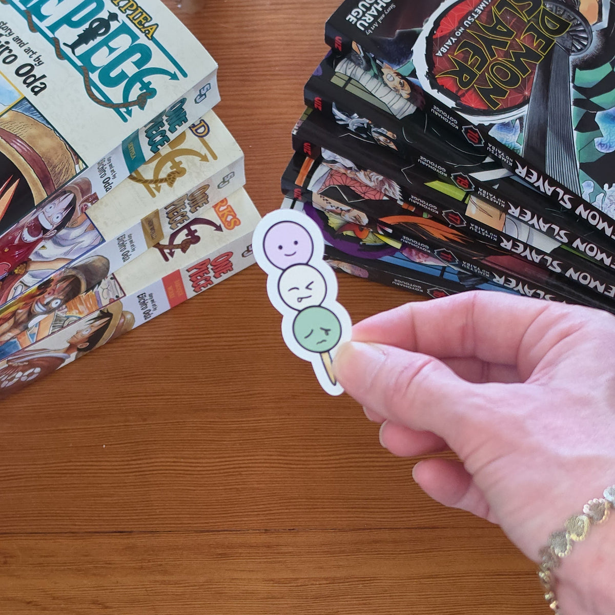A hand holds a die-cut sticker featuring three dango on a stick (pink, white, and light green) on a wooden table, with manga books in the background.