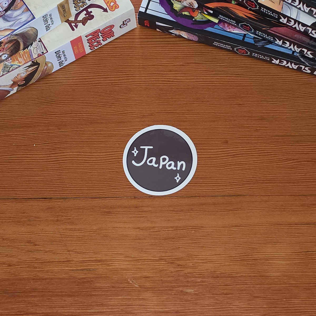 A round brown and white sticker with the word 'Japan' written on it, placed on a wooden table next to stacks of manga books.