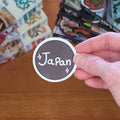 A hand holds a round sticker that says 'Japan' in white script on a dark brown background, with a wooden table and anime manga books in the blurred background.