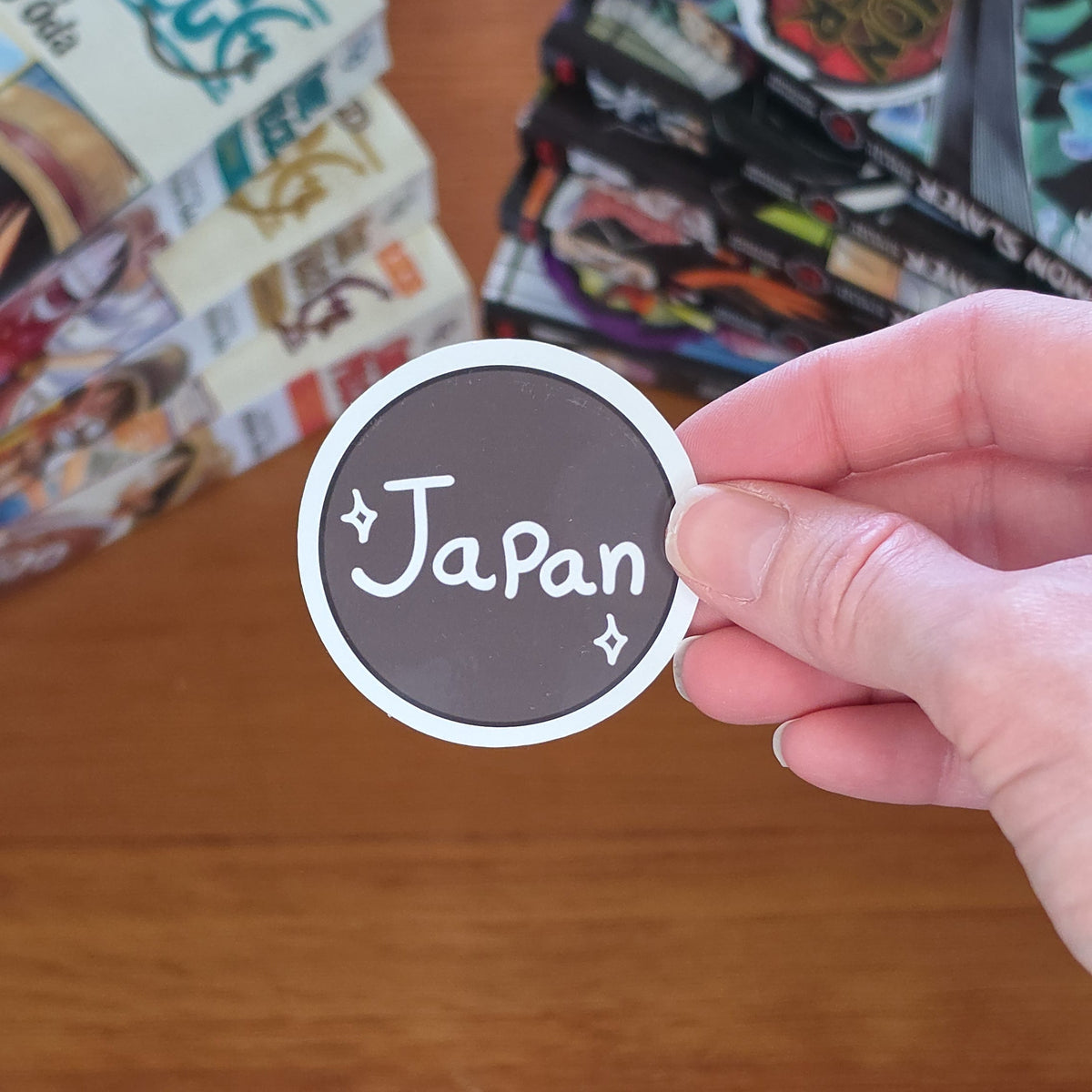 A hand holds a round sticker that says 'Japan' in white script on a dark brown background, with a wooden table and anime manga books in the blurred background.