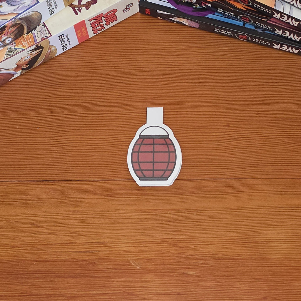 A small magnetic bookmark with a red paper lantern illustration with black grid lines, placed on a wooden tabletop. Manga books are stacked on both sides of the table.