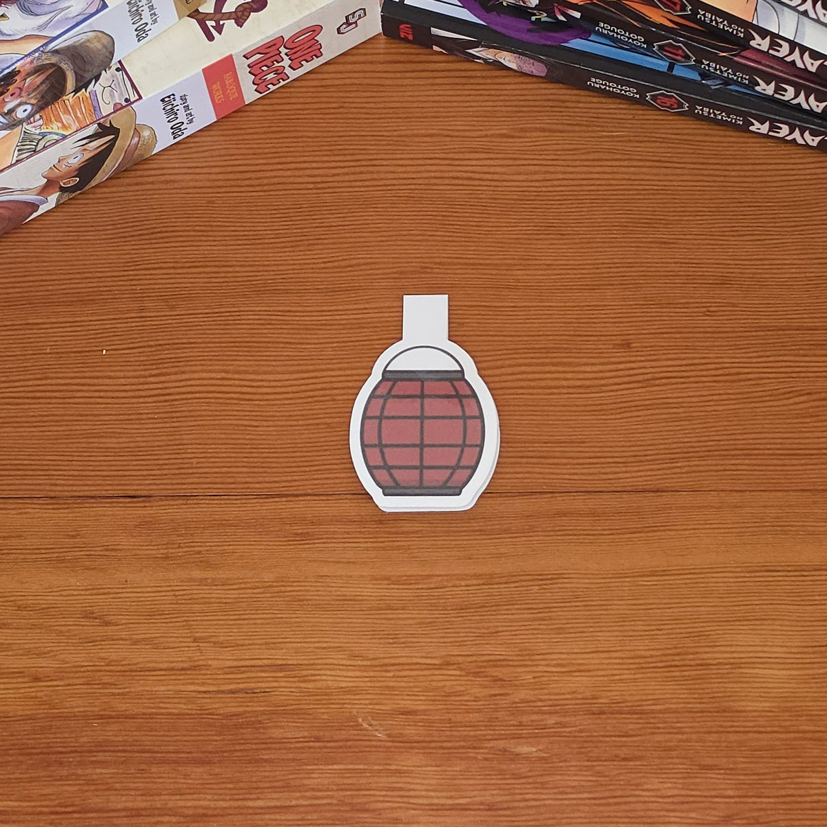 A small magnetic bookmark with a red paper lantern illustration with black grid lines, placed on a wooden tabletop. Manga books are stacked on both sides of the table.
