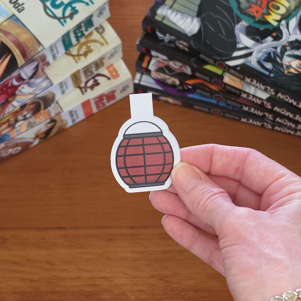 A hand holding a small magnetic bookmark with a red paper lantern illustration with black grid lines, with a wooden tabletop and stacks of manga books in the background.