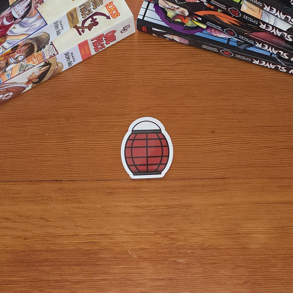 A small die-cut sticker shaped like a red paper lantern with a white border sits on a wooden tabletop. Manga books are stacked on both sides of the table.