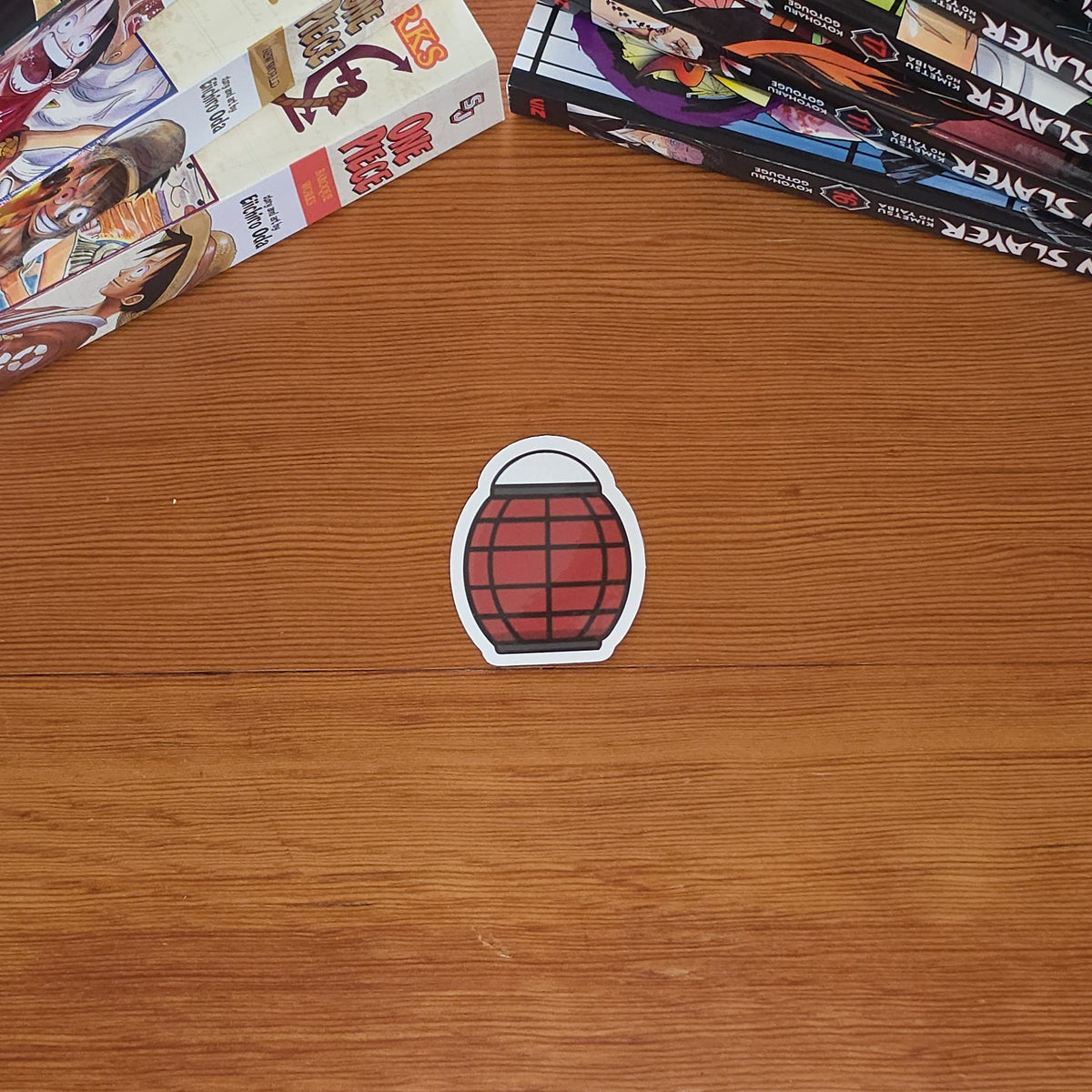 A small die-cut sticker shaped like a red paper lantern with a white border sits on a wooden tabletop. Manga books are stacked on both sides of the table.