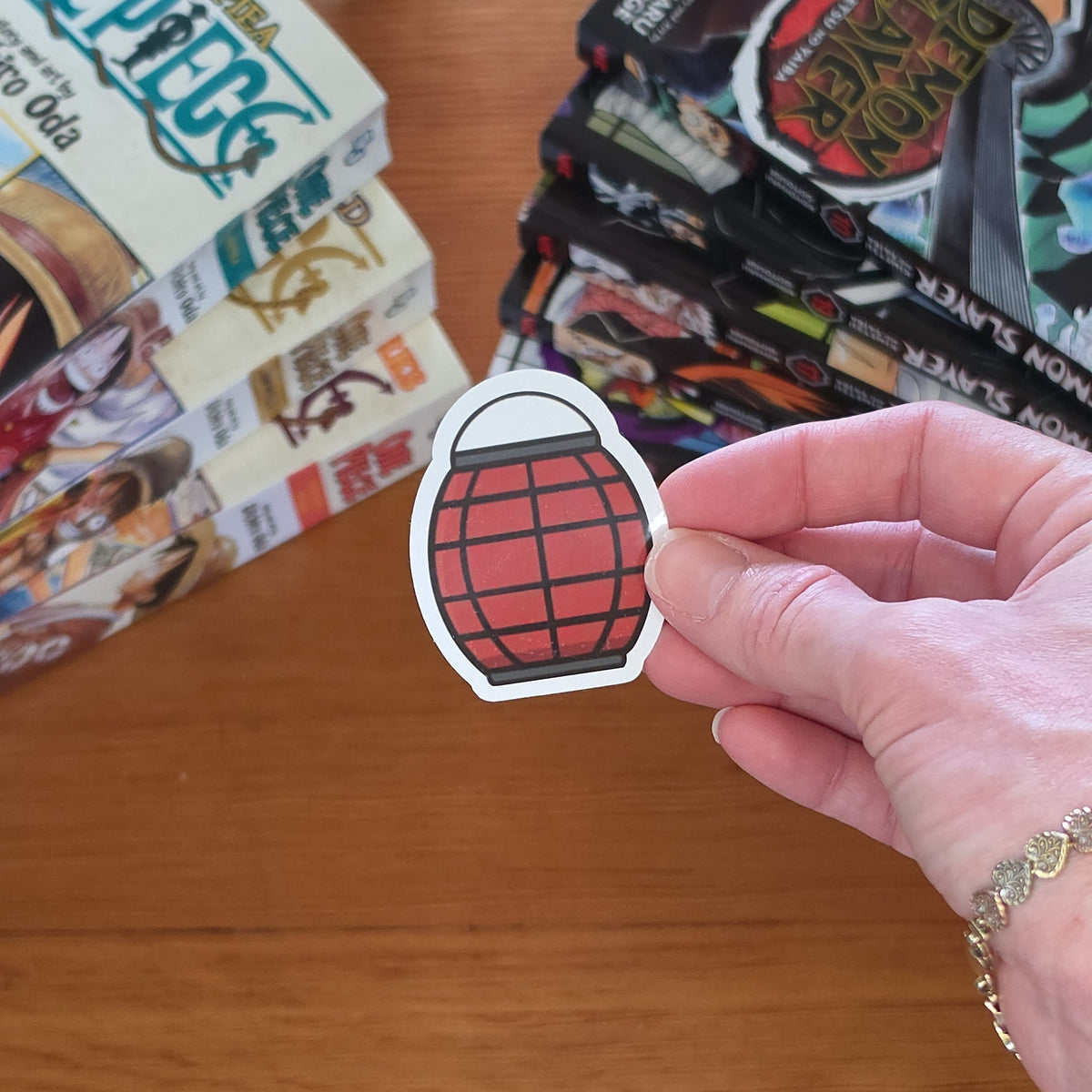 A hand holds a die-cut sticker shaped like a red paper lantern with black lines and a white border, set against a wooden tabletop with manga books in the background.