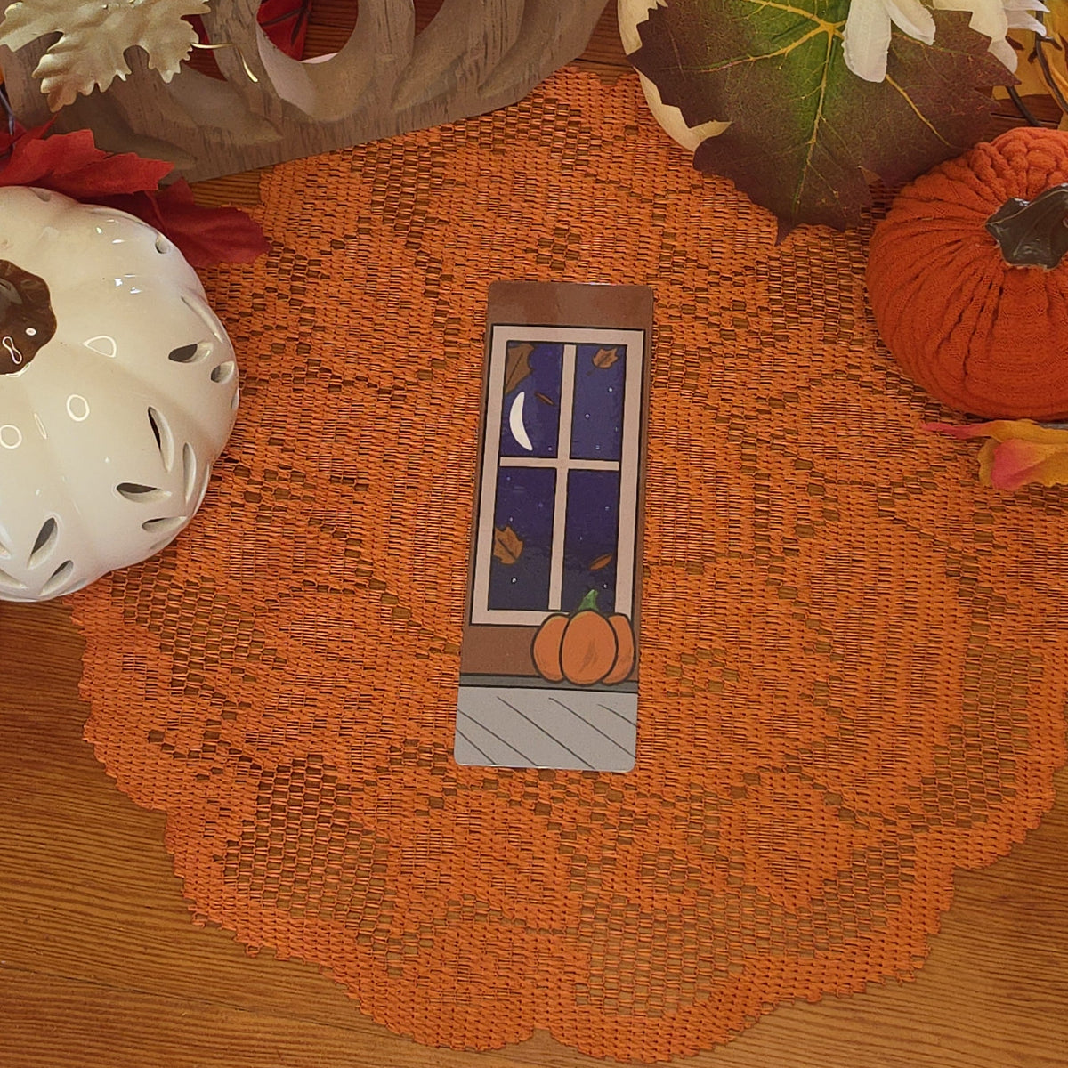 A rectangular bookmark featuring a pumpkin sitting on the ground near a window with the moon shining on it illustration, set against a cozy autumn-themed background with a lace doily, fall leaves, and pumpkin pottery.