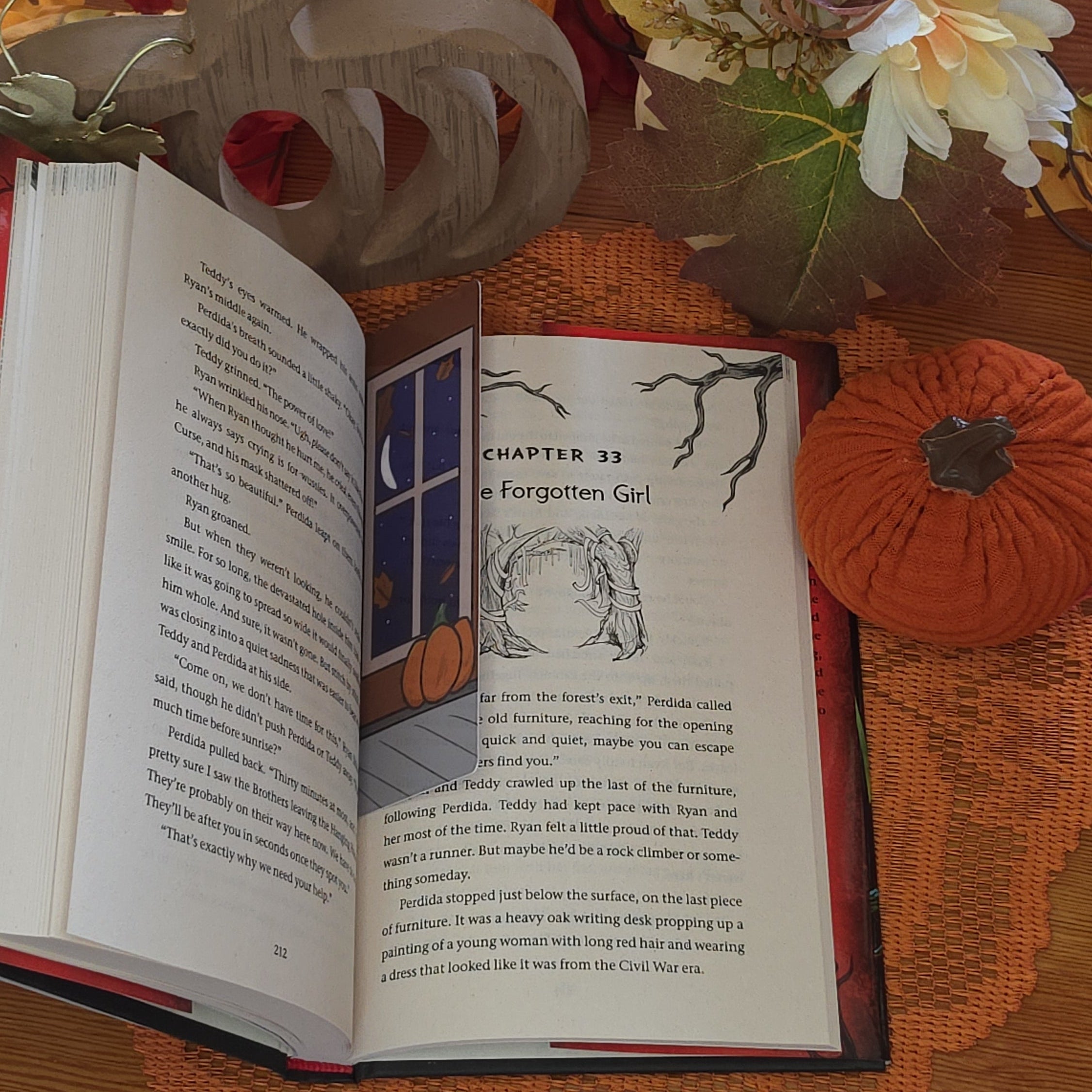 An open book features a rectangular bookmark featuring a pumpkin sitting on the ground near a window with the moon shining on it illustration, set against a cozy autumn-themed background with a lace doily, fall leaves, and pumpkin pottery.