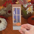 A hand holding a rectangular bookmark featuring a pumpkin sitting on the ground near a window with the moon shining on it illustration, set against a cozy autumn-themed background with a lace doily, fall leaves, and pumpkin pottery.
