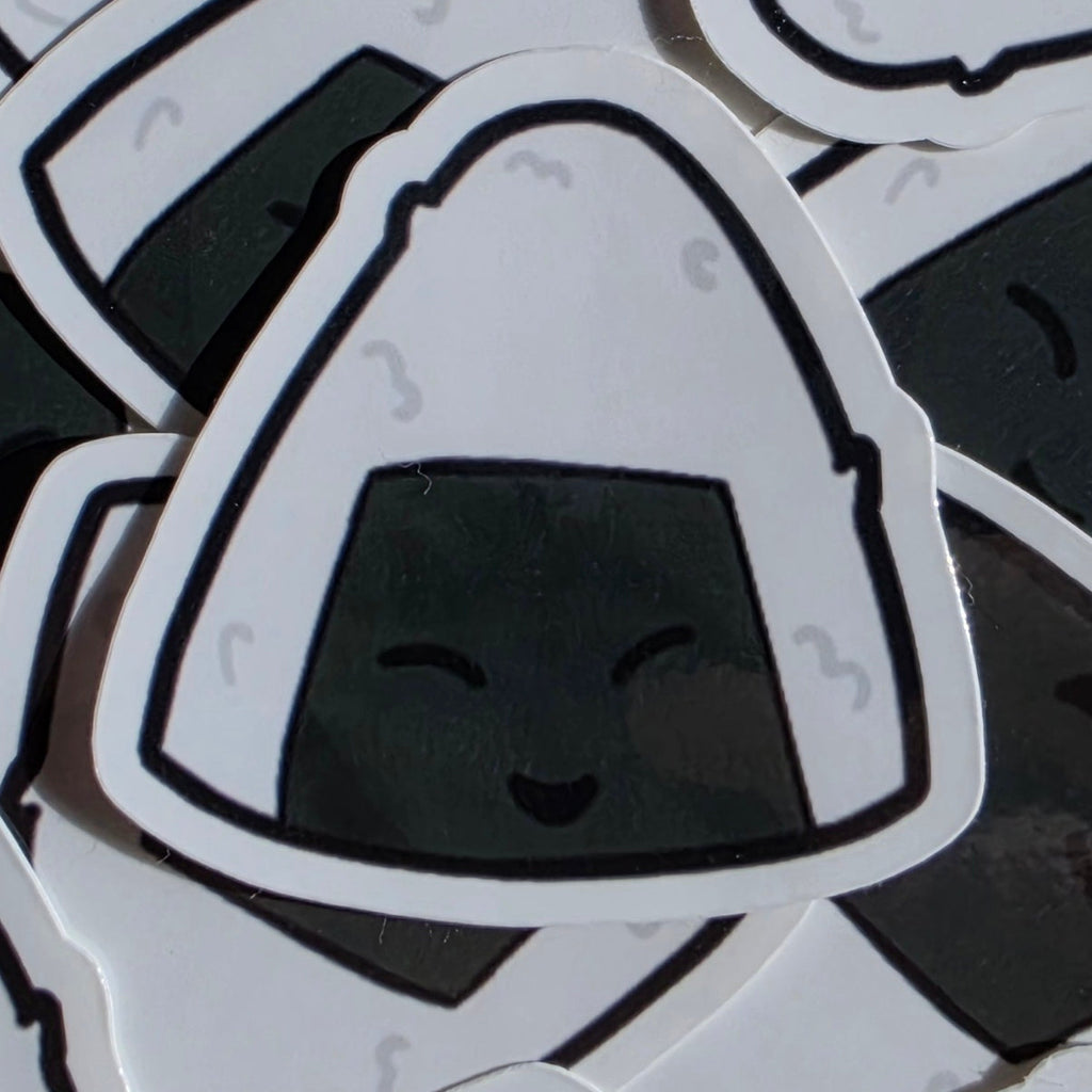 A close-up of several die-cut stickers shaped like onigiri (rice balls), arranged overlapping. The central sticker is white with a black outline and a dark gray smiling face, while the surrounding stickers show the same design from different angles.