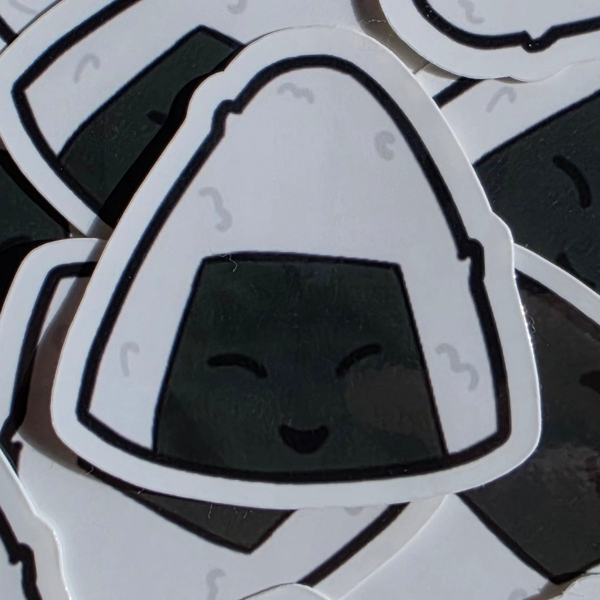 A close-up of several die-cut stickers shaped like onigiri (rice balls), arranged overlapping. The central sticker is white with a black outline and a dark gray smiling face, while the surrounding stickers show the same design from different angles.