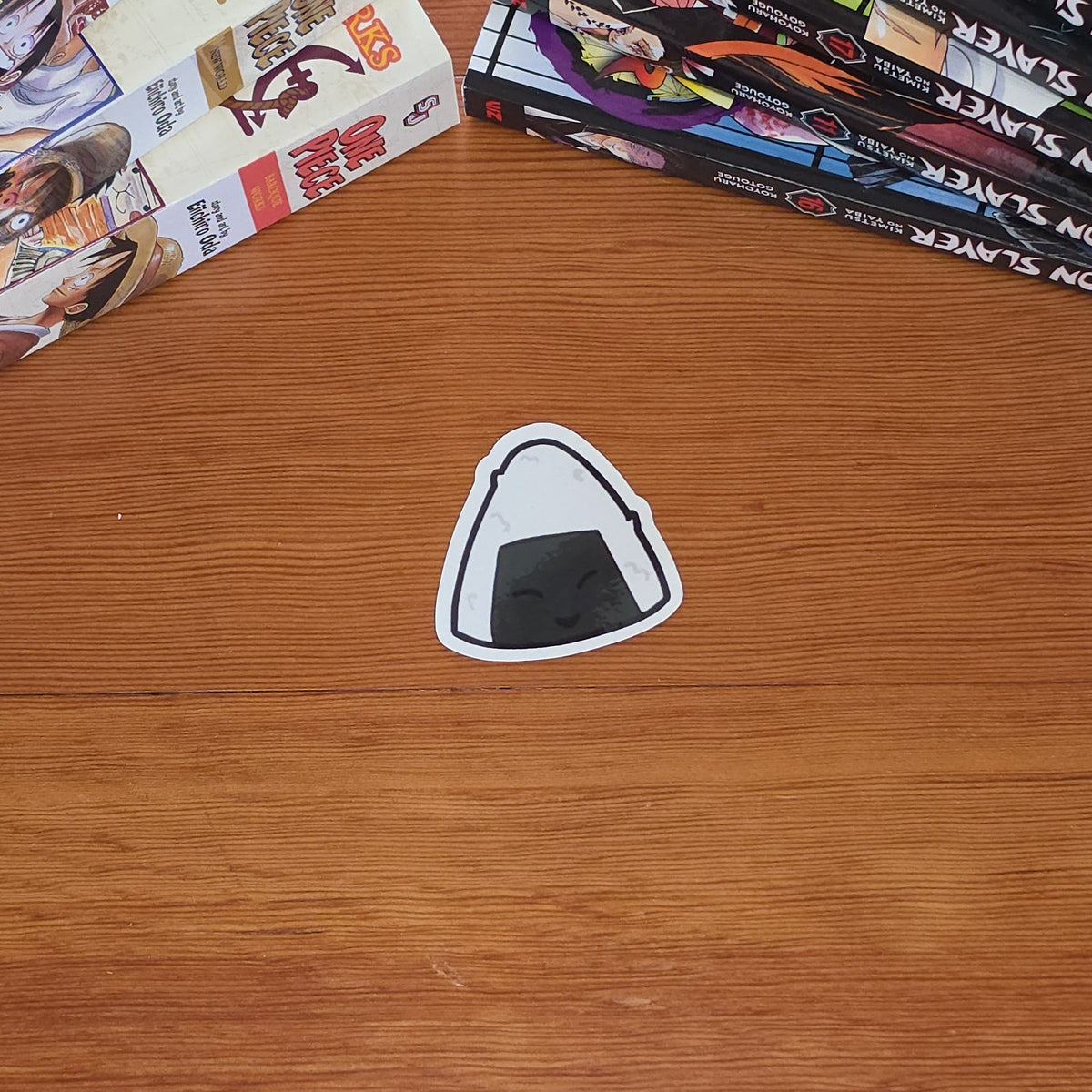 A small triangular sticker with a black-and-white riceball (onigiri) style graphic sits on a wooden tabletop. Stacks of manga books are visible along the top left and right edges of the frame.