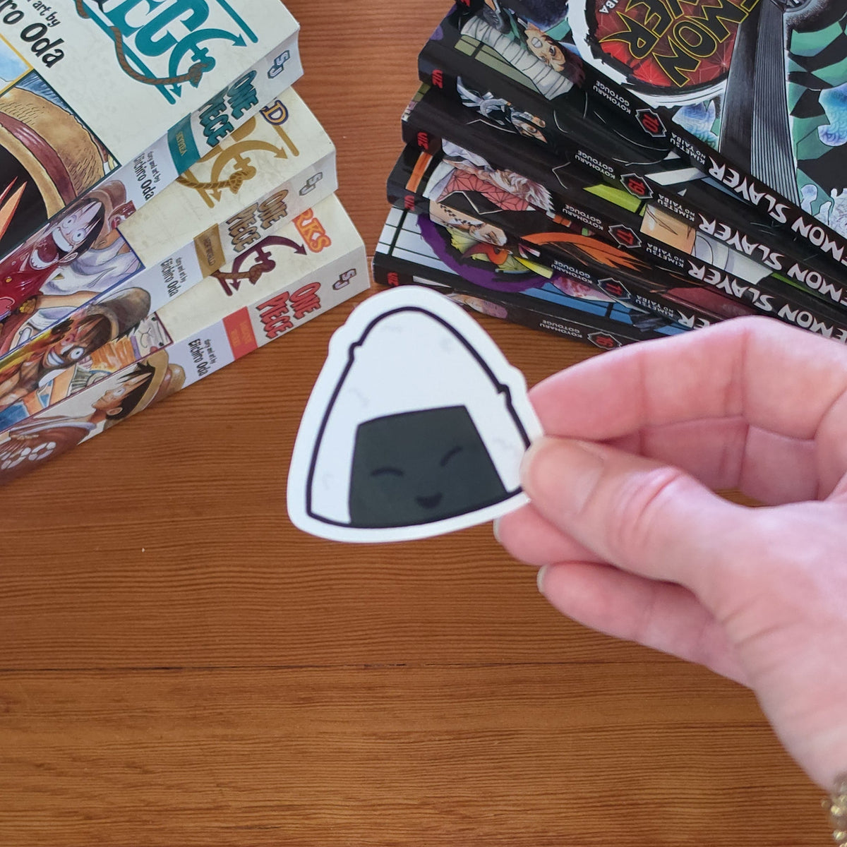 A hand holding a small die-cut sticker shaped like an onigiri rice ball, with a wooden tabletop and manga books in the background.