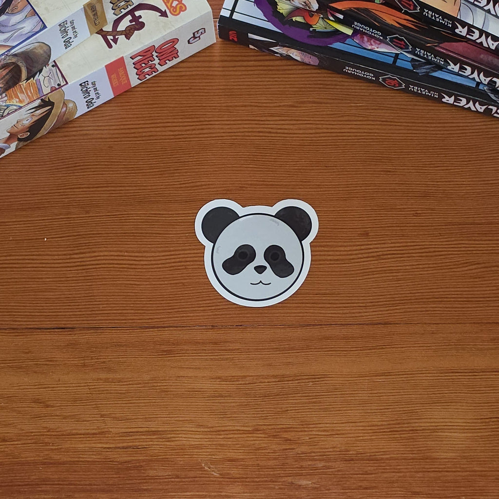 A small panda-shaped sticker sits on a wooden tabletop, with stacks of manga books visible along the top left and right edges.