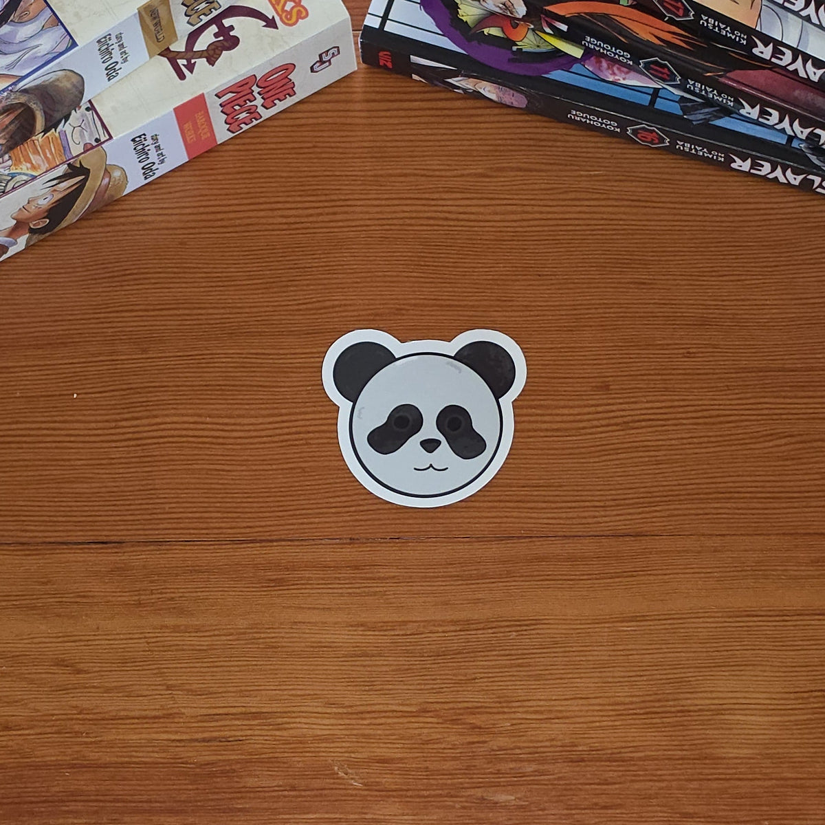 A small panda-shaped sticker sits on a wooden tabletop, with stacks of manga books visible along the top left and right edges.
