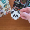 A hand holds a die-cut sticker shaped like a panda face with black and grey coloring, set against a wooden tabletop with manga books in the background.