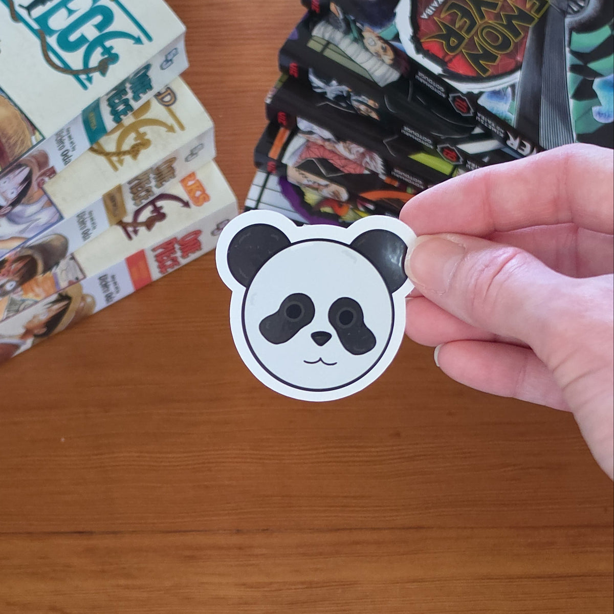 A hand holds a die-cut sticker shaped like a panda face with black and grey coloring, set against a wooden tabletop with manga books in the background.