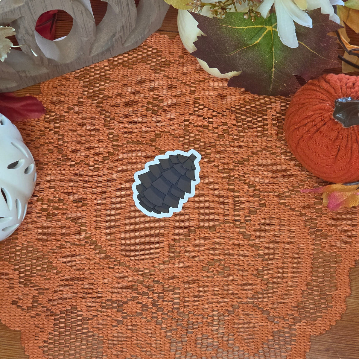 A sticker in the shape of a pinecone sitting on an orange doily with autumn decor in the background.