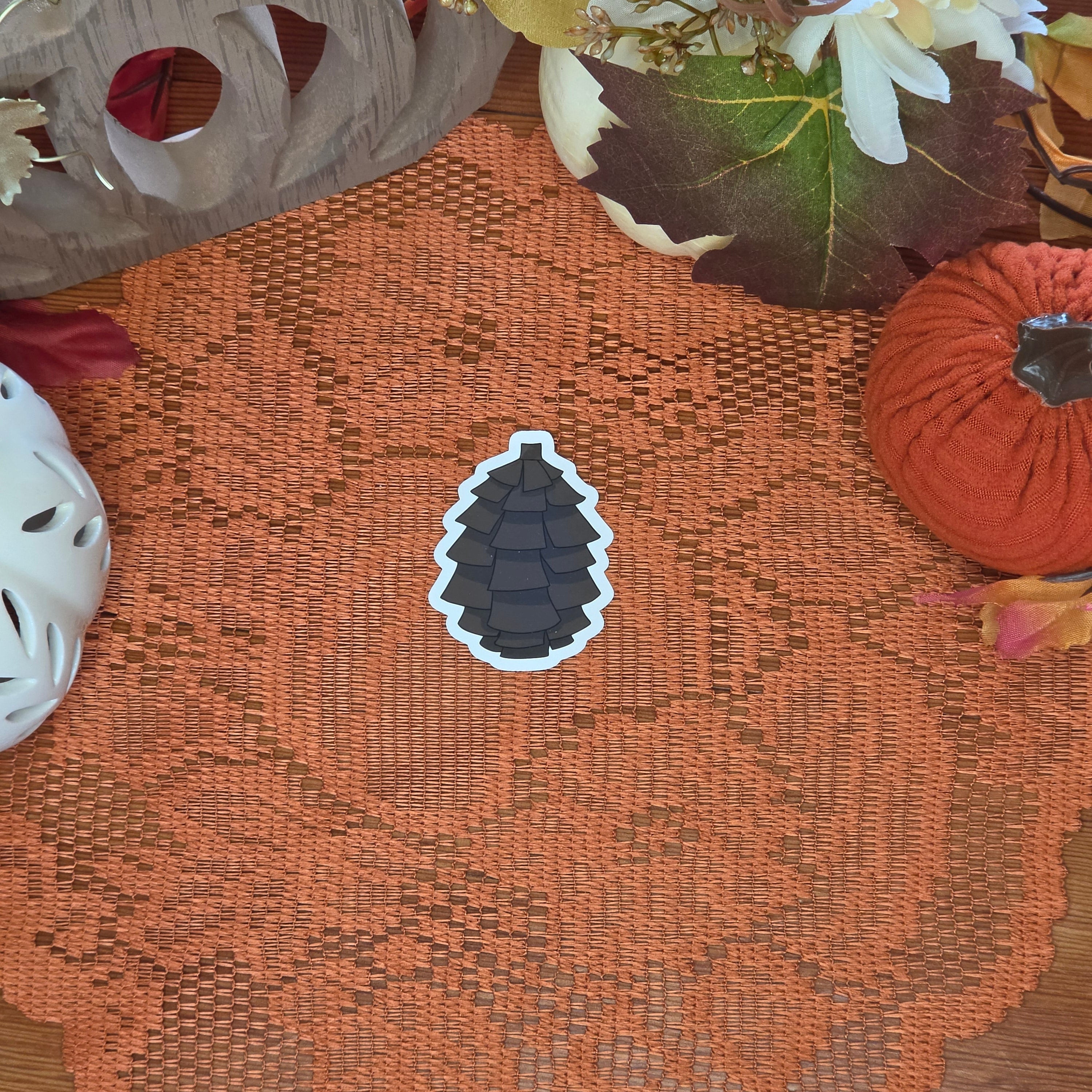 A sticker in the shape of a pinecone sitting on an orange doily with autumn decor in the background.