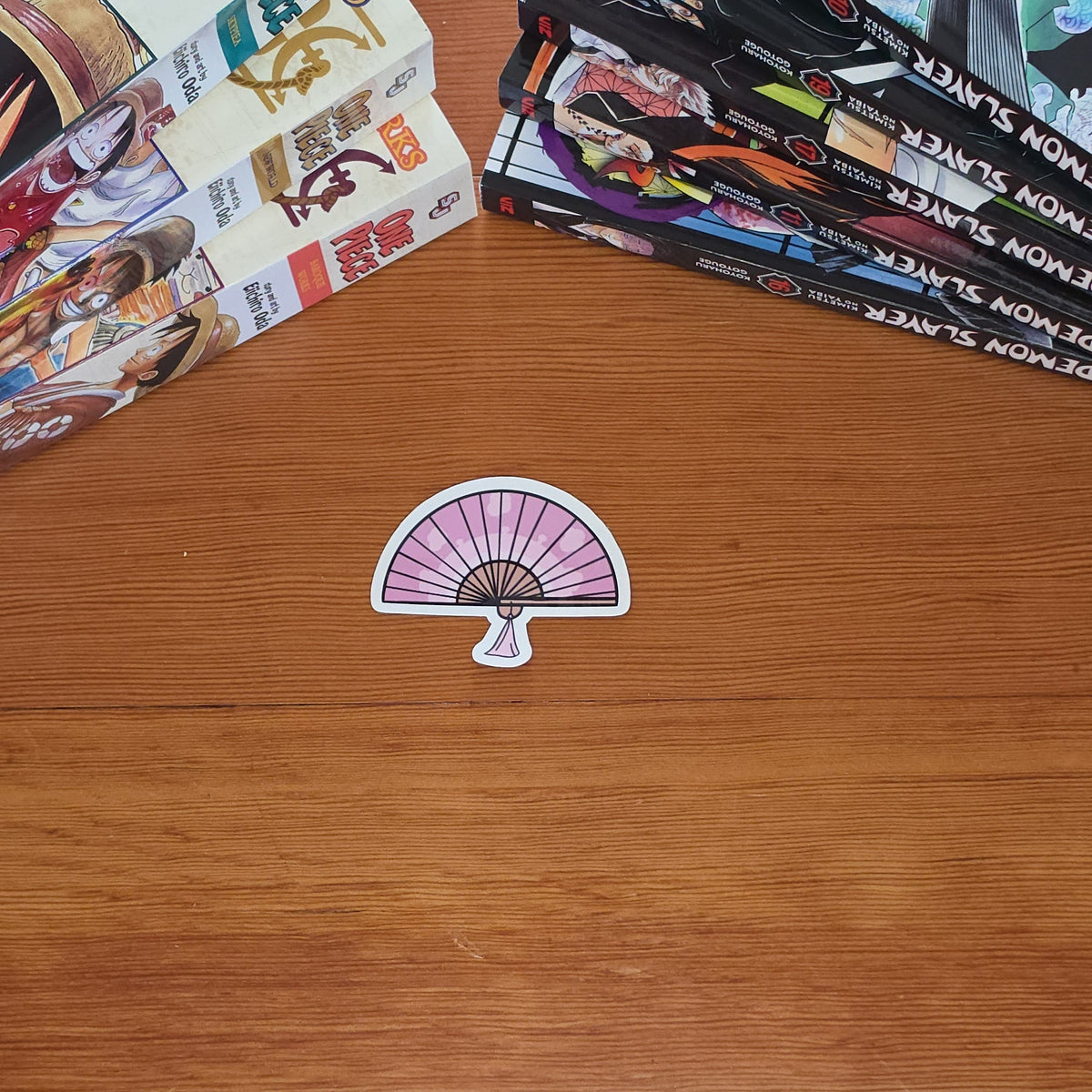 A small die-cut sticker shaped like a pink and white folding fan with black lines, placed on a wooden tabletop. Stacks of manga books are visible in the background on both sides.