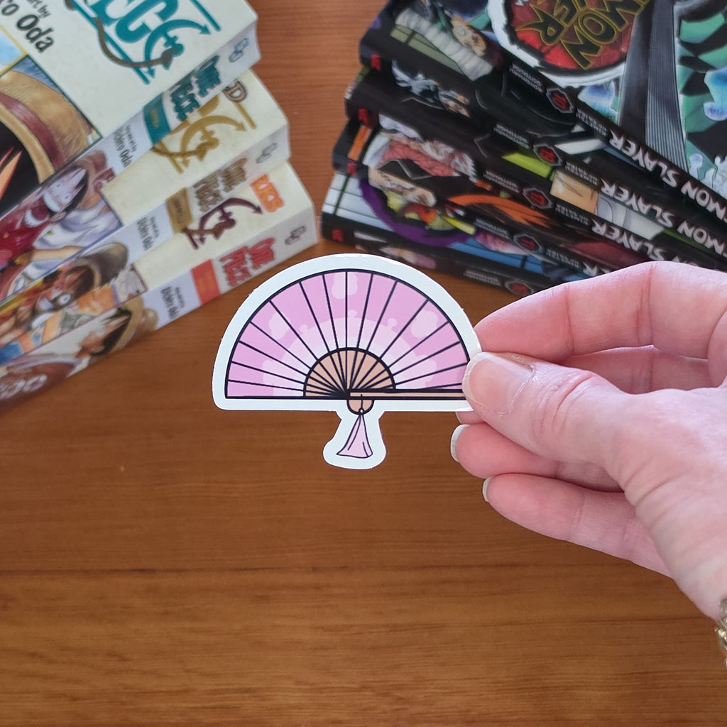 A hand holds a die-cut sticker shaped like a pink and white folding fan with black line details, set against a wooden tabletop with manga books in the background.