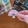 A hand holds a die-cut sticker shaped like a pink and white folding fan with black line details, set against a wooden tabletop with manga books in the background.