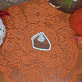 A sticker in the shape of a slice of pumpkin pie with whipped cream topping sitting on an orange doily with autumn decor in the background.