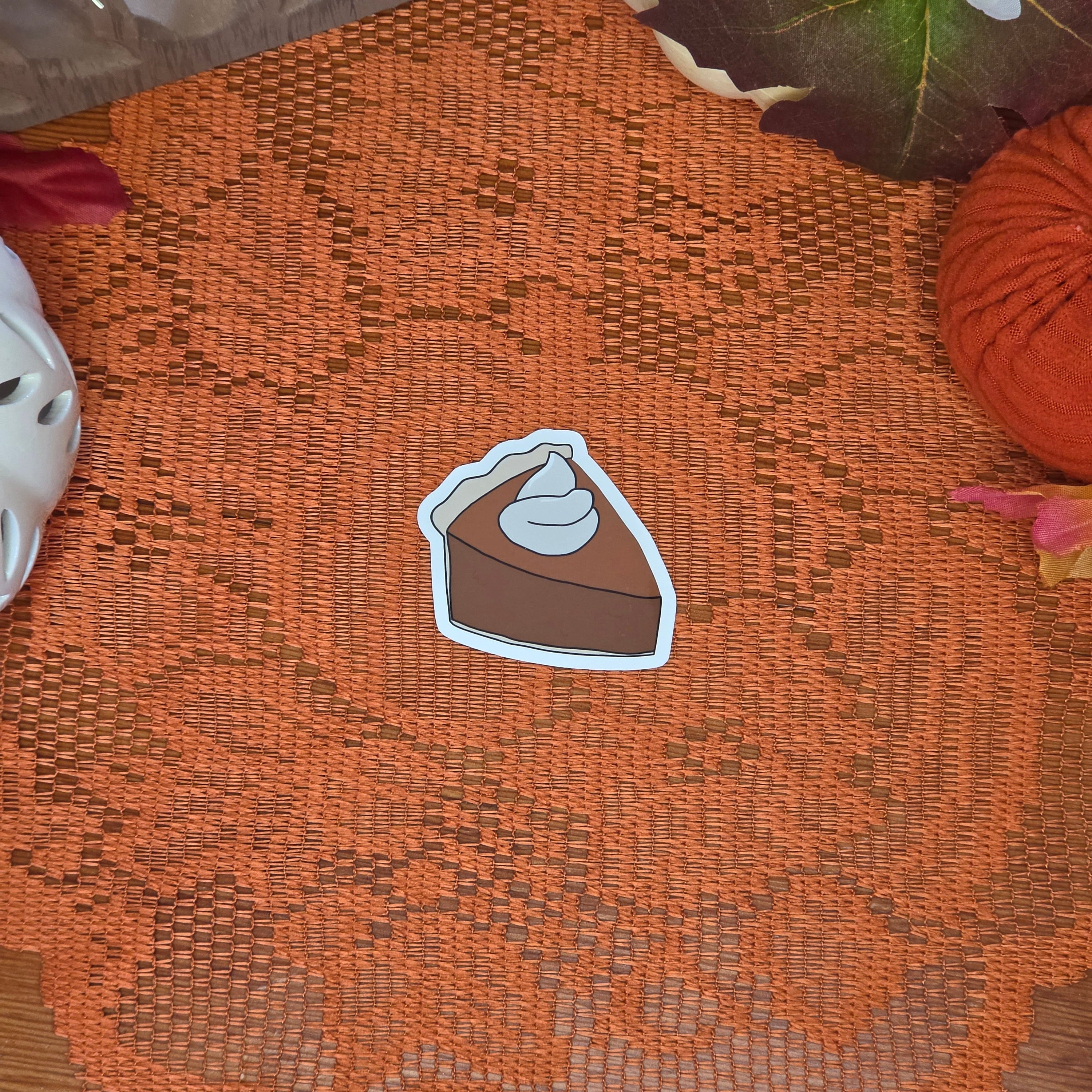 A sticker in the shape of a slice of pumpkin pie with whipped cream topping sitting on an orange doily with autumn decor in the background.