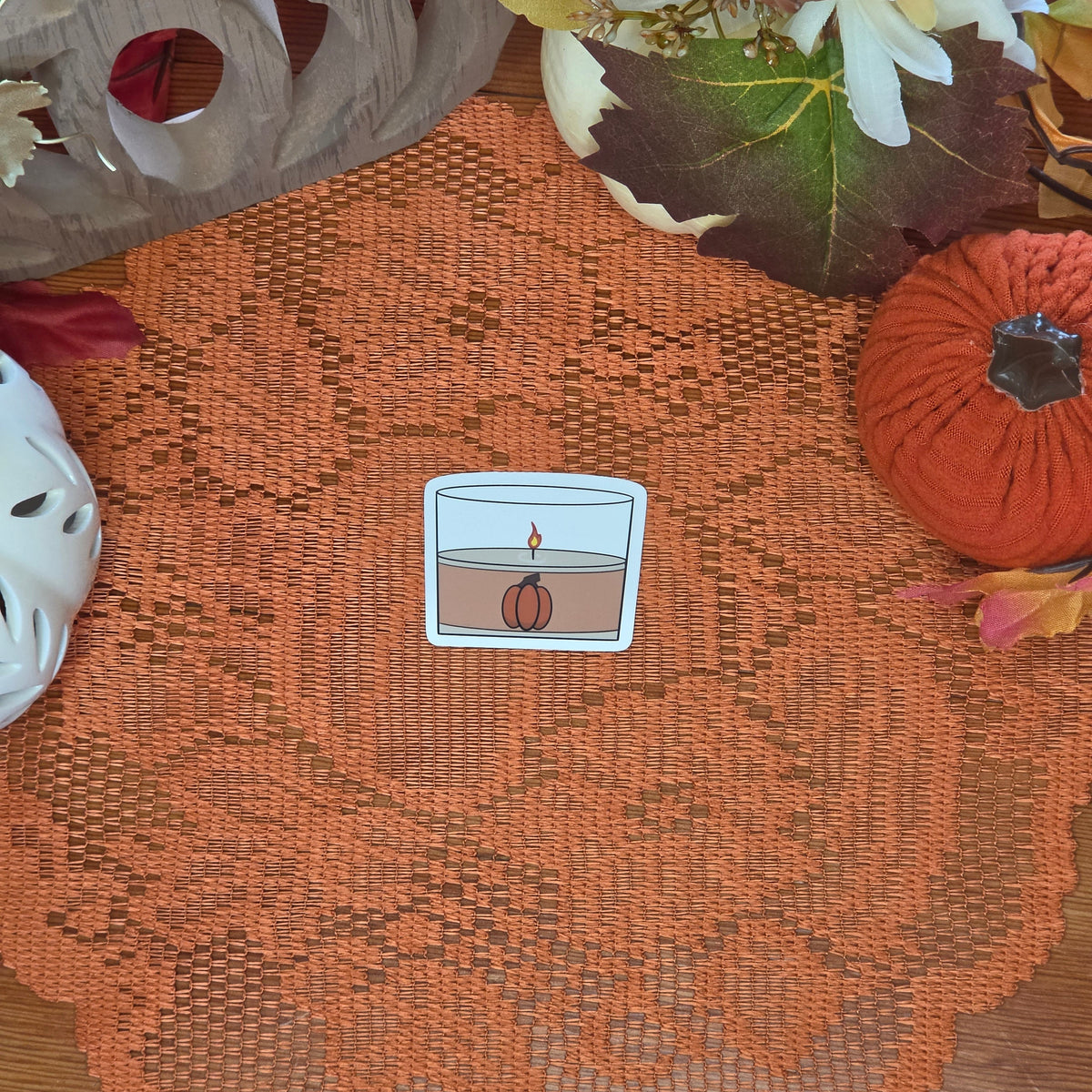 A sticker in the shape of a pumpkin spice candle sitting on an orange doily with autumn decor in the background.