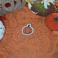 A sticker in the shape of a pumpkin sitting on an orange doily with autumn decor in the background.