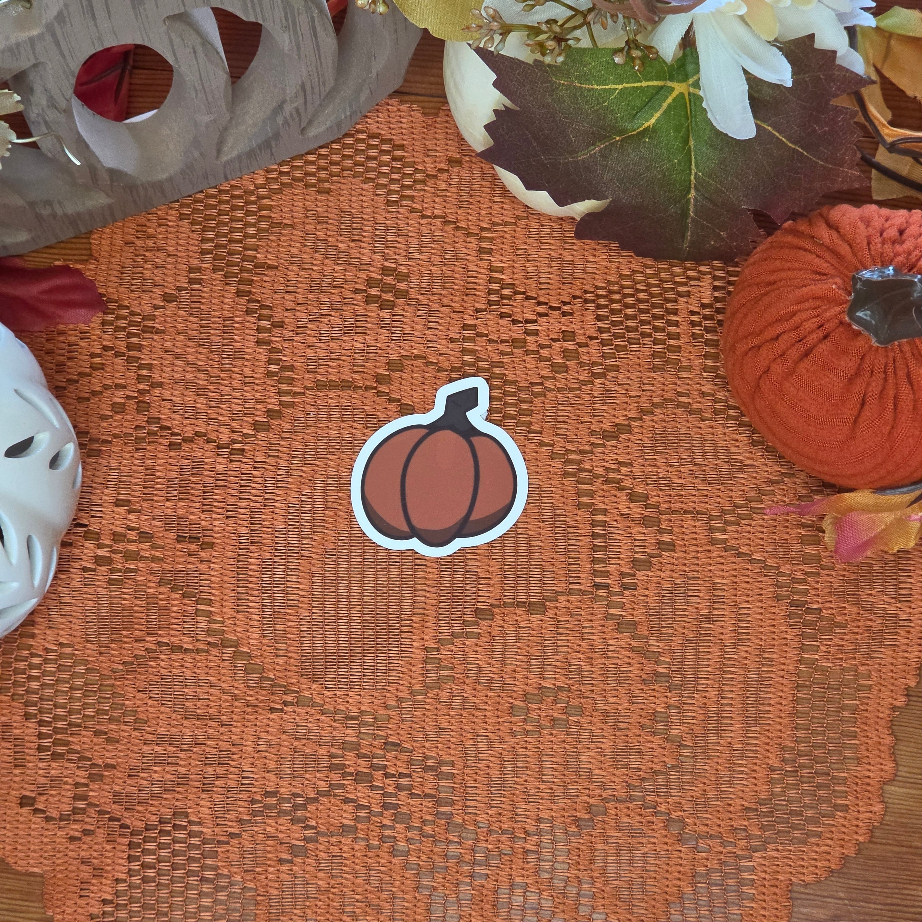 A sticker in the shape of a pumpkin sitting on an orange doily with autumn decor in the background.