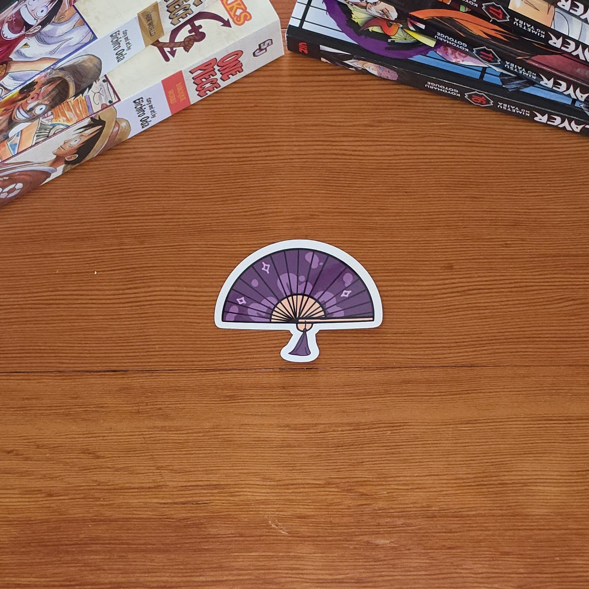A die-cut sticker featuring a purple hand fan design with beige ribs and light gray outlines, set against a wood table with stacks of manga books in the background.