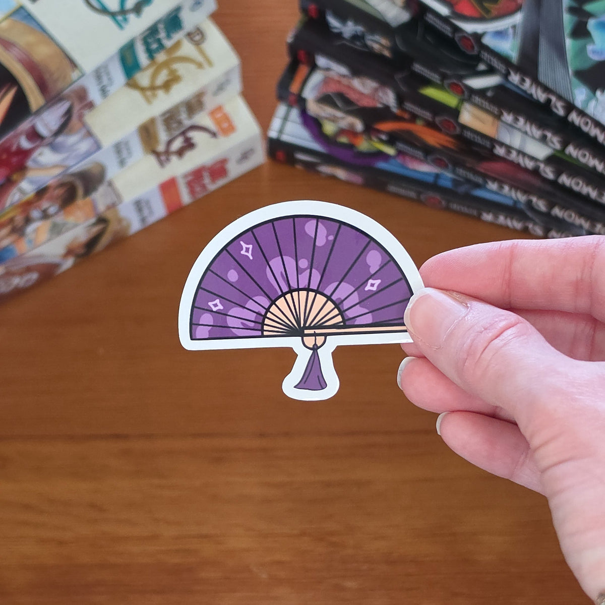A hand holding a die-cut sticker featuring a purple hand fan design with beige ribs and light gray outlines, set against a wood table with stacks of manga books in the background.