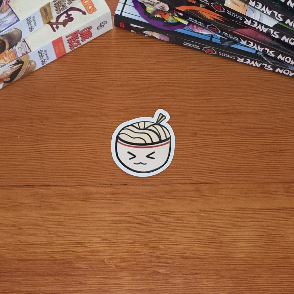 A small die-cut sticker featuring a cute cartoon ramen bowl with a smiling face, placed on a wooden tabletop next to stacks of manga books.