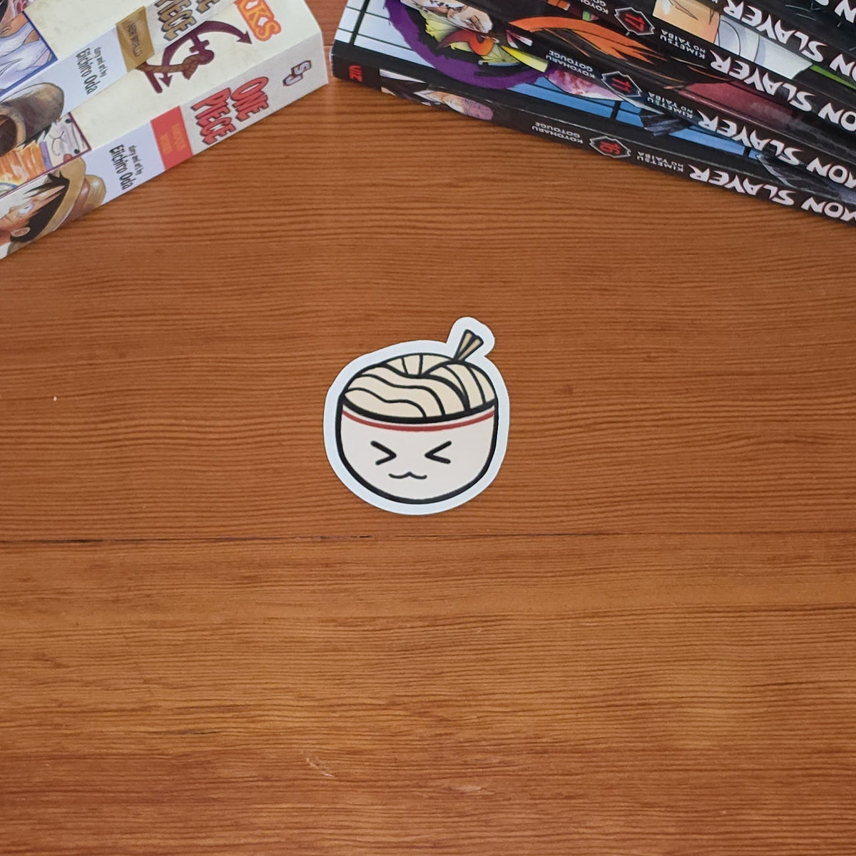 A small die-cut sticker featuring a cute cartoon ramen bowl with a smiling face, placed on a wooden tabletop next to stacks of manga books.