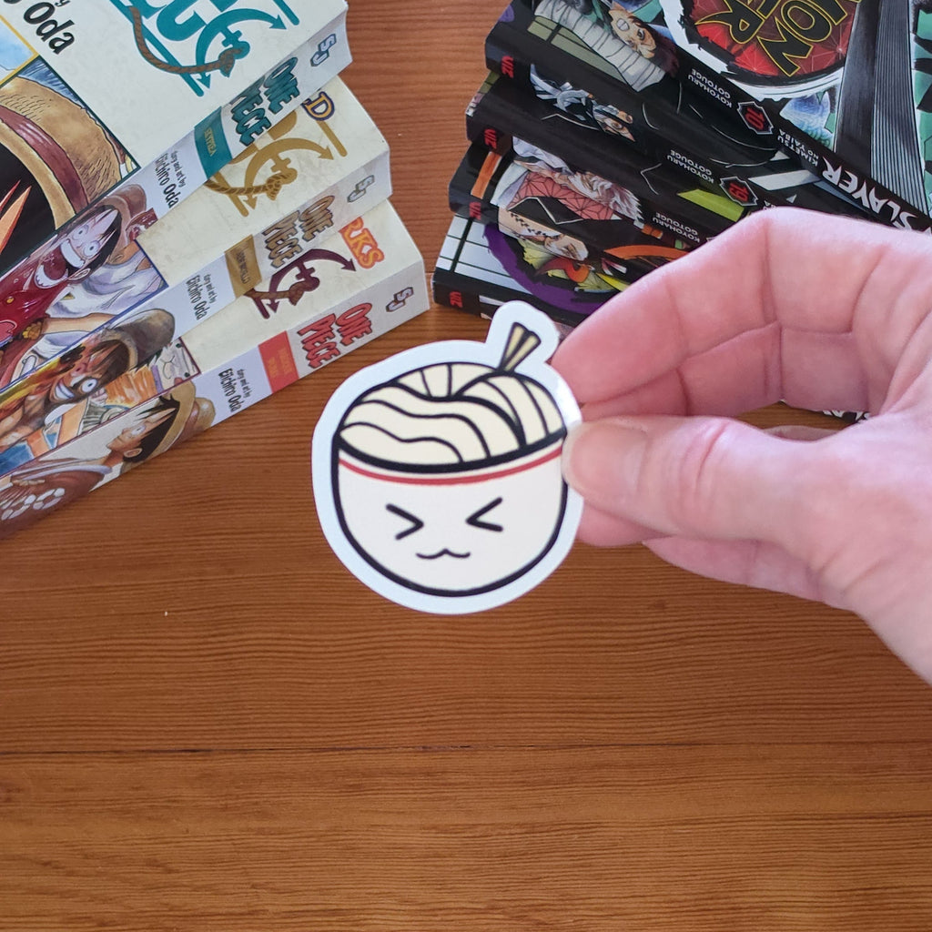 A hand holds a round sticker featuring a cartoon ramen bowl with a winking face, set against a wooden tabletop with manga books in the background.