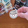 A hand holds a round sticker featuring a cartoon ramen bowl with a winking face, set against a wooden tabletop with manga books in the background.