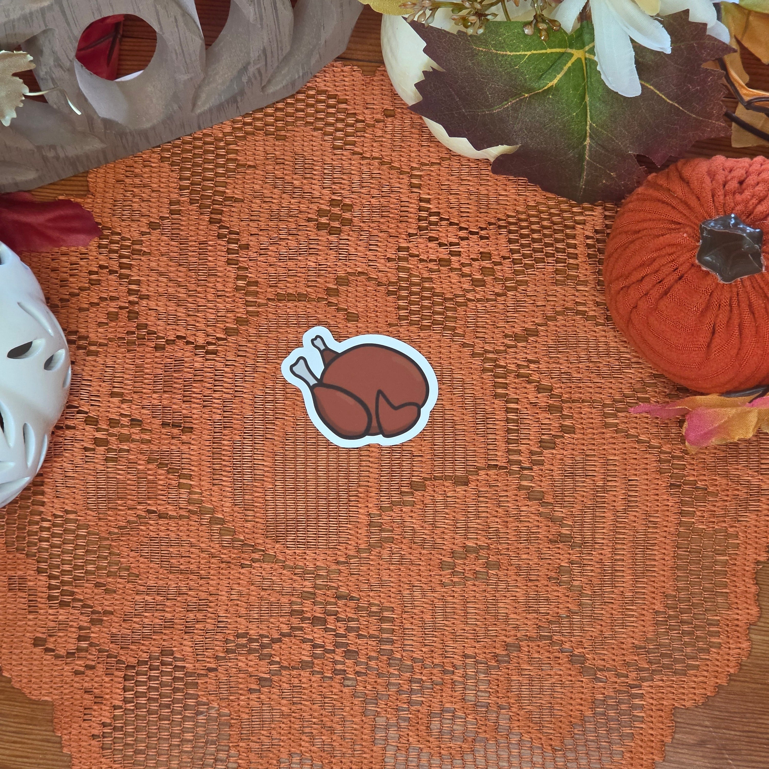 A sticker in the shape of a roast turkey sitting on an orange doily with autumn decor in the background.