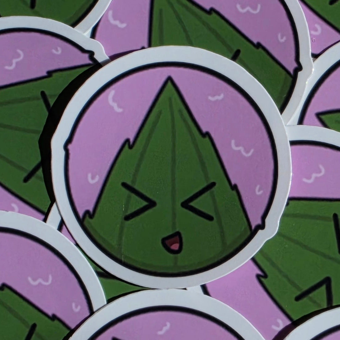 A close-up of round stickers featuring a green leaf wrapped rice ball with a character face inside a lavender sakuramochi ball with black outlines and white borders, with additional stickers partially visible around it.