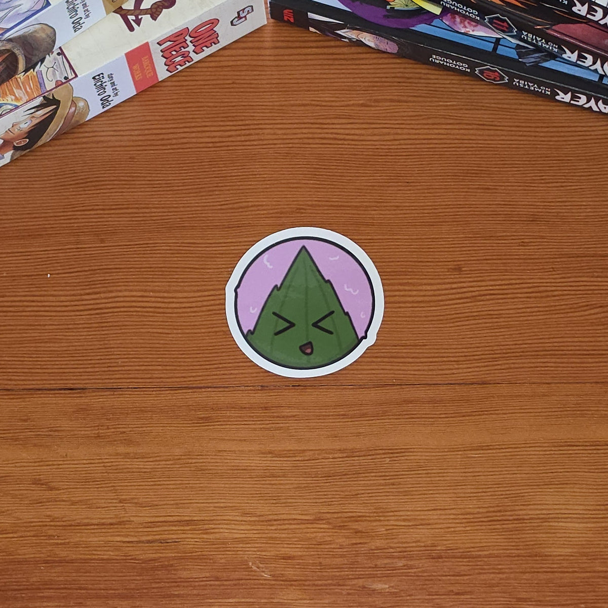A round sticker featuring a green leaf wrapped rice ball with a character face inside a lavender sakuramochi ball with black outlines and white border placed on a wooden tabletop next to stacks of manga books.
