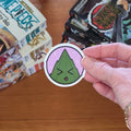 A hand holds a round sticker featuring a green leaf wrapped rice ball with a character face inside a lavender sakuramochi ball with black outlines and white borders, set against a wooden table with manga books in the background.