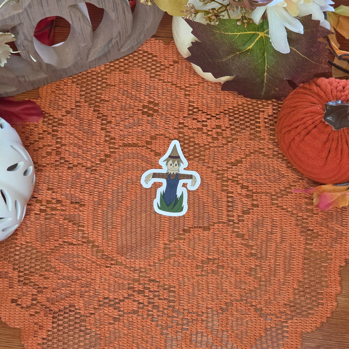A sticker in the shape of a scarecrow sitting on an orange doily with autumn decor in the background.