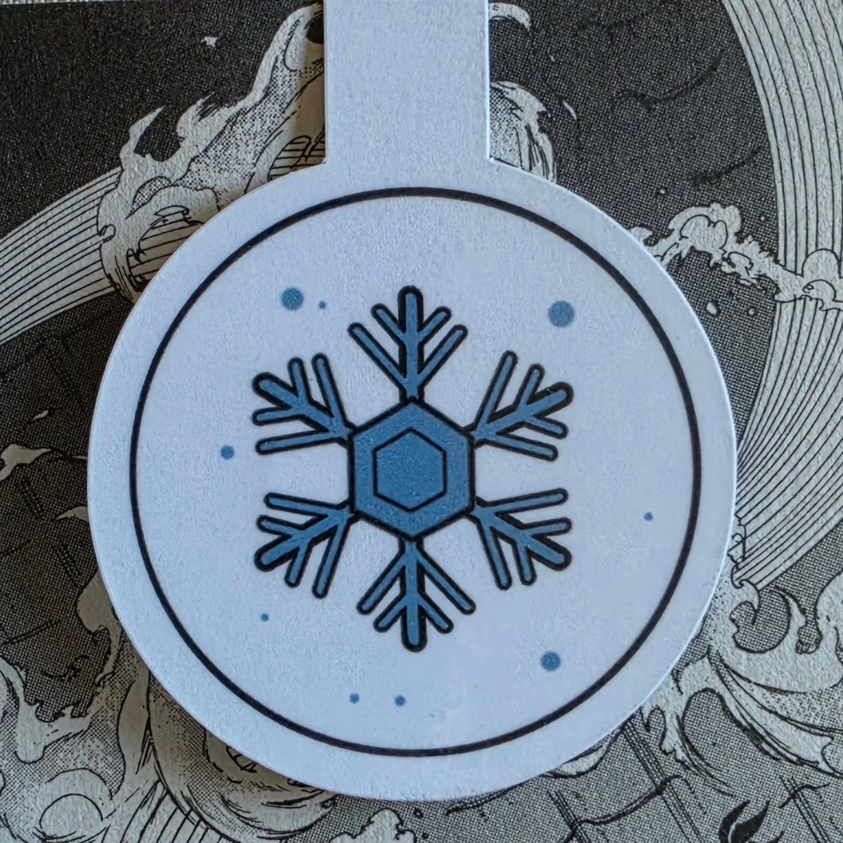 A close-up of a round white magnetic bookmark with a blue snowflake design and black circular border, placed over a gray manga illustration background with wave-like patterns.