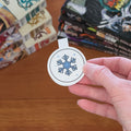 A hand holding a round white sticker featuring a blue snowflake design, with stacks of manga books on a wooden table in the background.