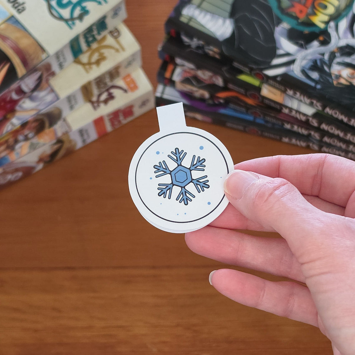 A hand holding a round white sticker featuring a blue snowflake design, with stacks of manga books on a wooden table in the background.