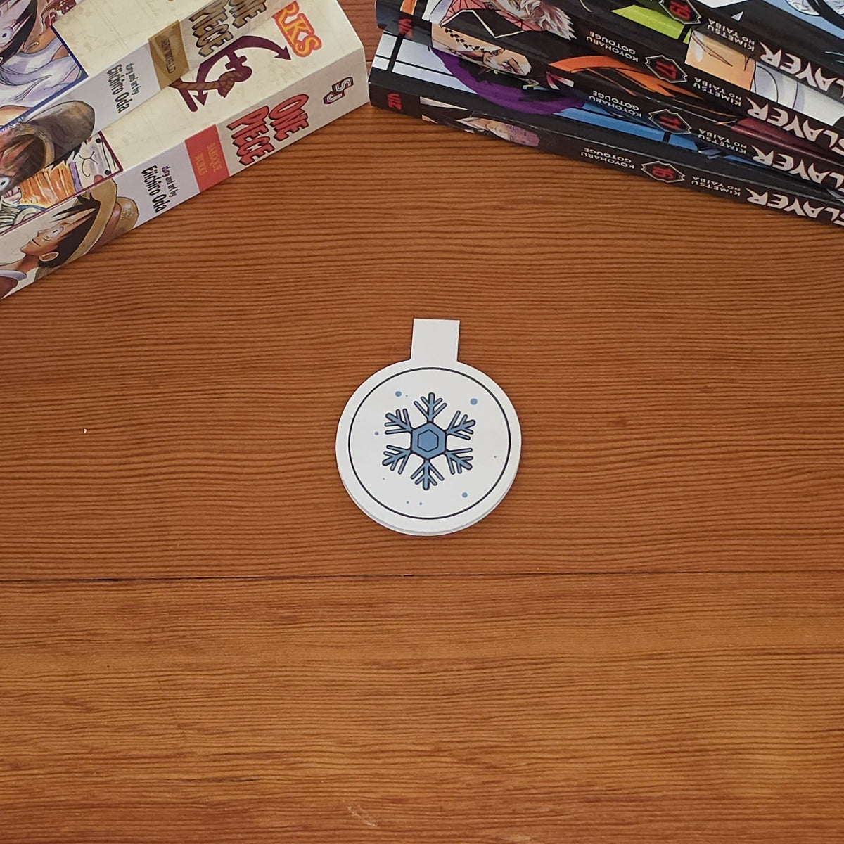 A small round white magnetic bookmark with a blue snowflake design sits on a wooden tabletop. Manga books are stacked on both sides of the surface.