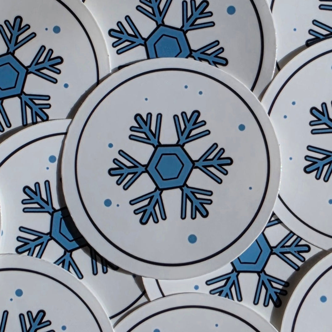 A close-up of round white stickers featuring a blue snowflake design with black outlines and small blue dots, arranged overlapping.