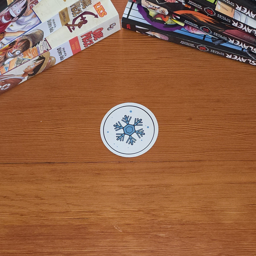 A small round white sticker with a blue snowflake sits on a wooden tabletop, with stacks of manga books visible in the top-left and top-right corners.