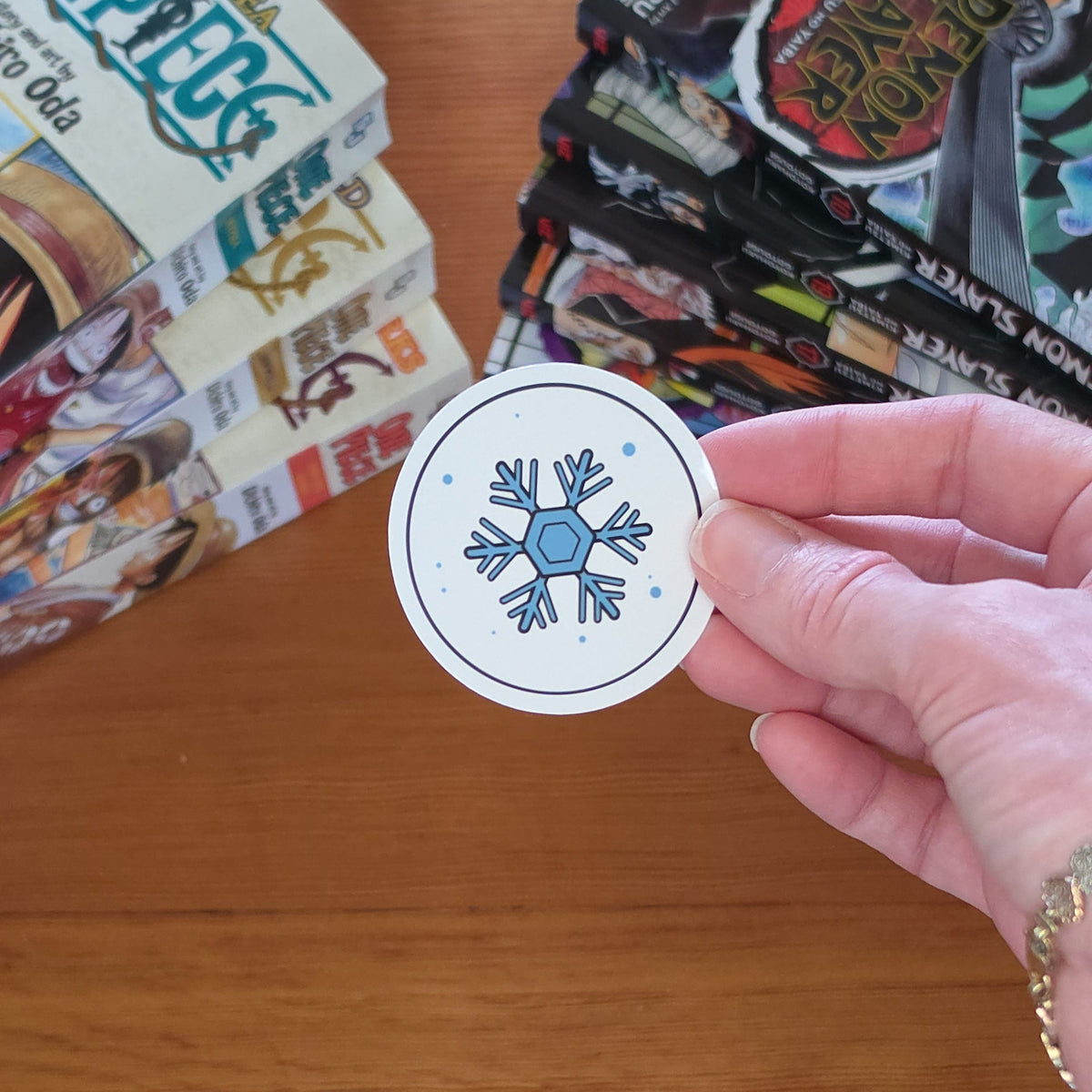 A hand holding a round white sticker with a blue snowflake design, set against a wooden table background with manga books.